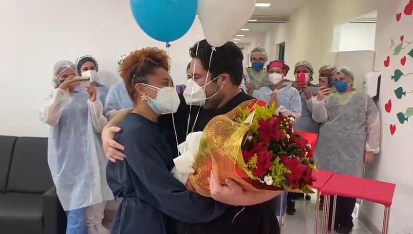 Homem com buque de rosas abraça namorada. Ao fundo vários enfermeiros tirando foto. #paratodosverem