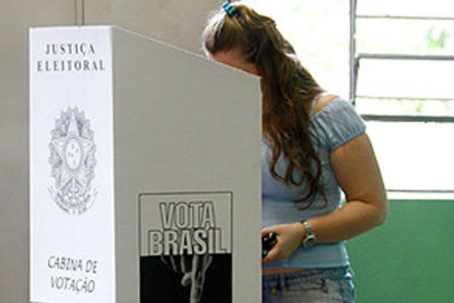 mulher vota atrás de cabine. Na cabine se justiça eleitoral, o brasão das armas nacional e cabine de votação. Na lateral, se lê Vota Brasil. Não se vê o rosto da mulher na cabine. #paratodosverem