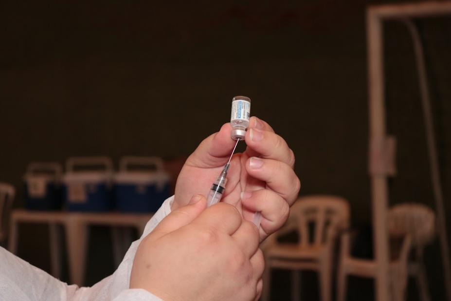 imagem em close sobre mãos preparando vacina. A mão esquerda segura o frasco de cabeça para baixo e a direita, a seringa com agulha, que entra no frasco. #paratodosverem