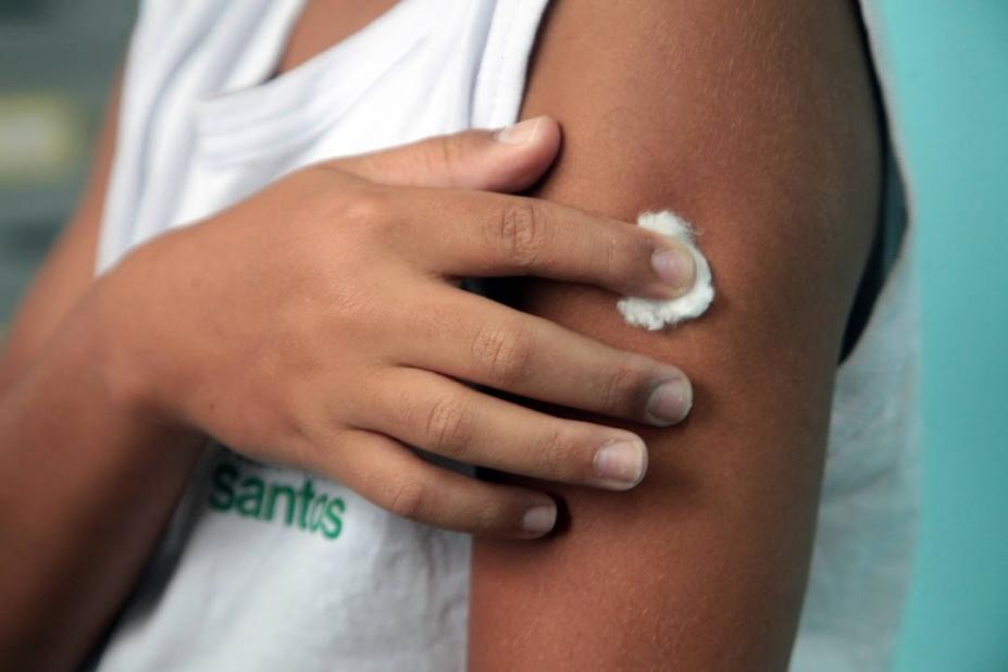 mão de criança uniformizada segura pedaço de algodão num braço após aplicação de vacina. #paratodosverem
