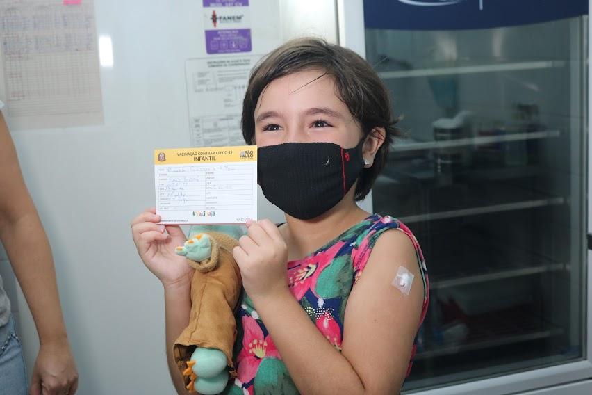 meninas segura comprovante de vacina. Ela tem um curativo no braço esquerdo. Ela também segura um bonequinho no outro braço. #paratodosverem
