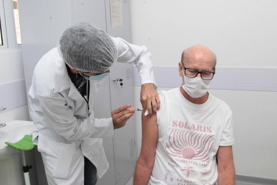 Homem está sentado em cadeira sendo vacinado por outro homem paramentado com avental, gorro e máscara. #Paratodosverem