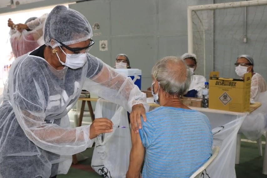 mulher está em pé usando touca, avental e máscara. Ela vacina homem que está sentado. #paratodosverem