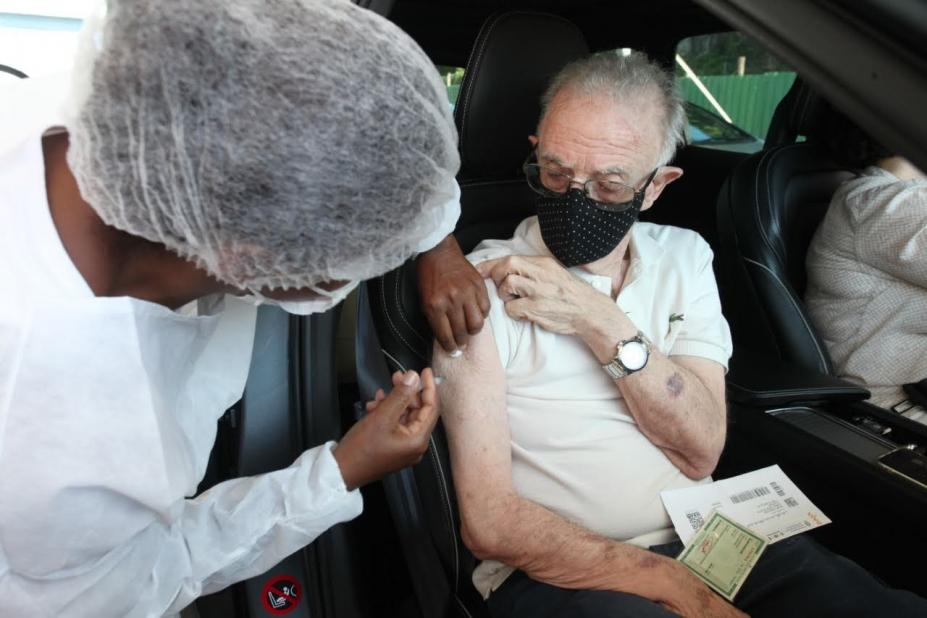 Mulher vestida de branco vacina idoso dentro de carro. Ele está segurando a manga da blusa e tem documentos no colo. #Paratodosverem