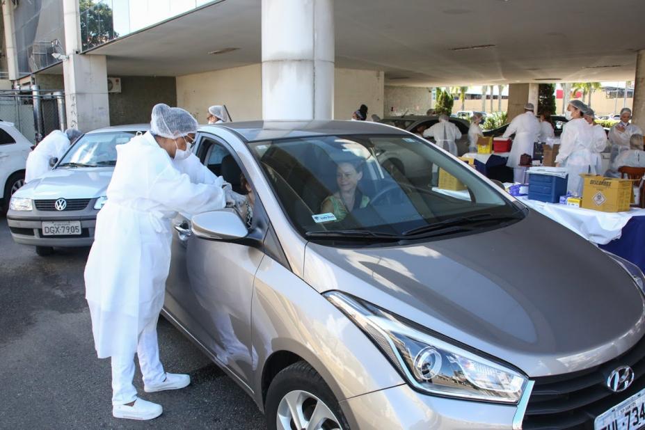 Mulher vestida de branco, avental, luvas, touca e máscara vacina alguém no banco do passageiro do carro parado. #Paratodosverem