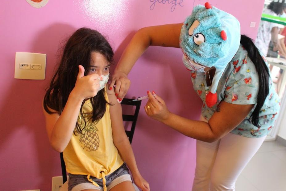 menina está sentada tomando vacina no braço. Ela faz sinal de positivo com a mão direita. Uma mulher com um chapéu colorida aplica a vacina. #paratodosverem