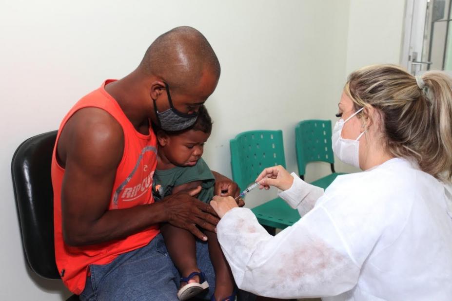 Homem segura uma criança que vai tomar a vacina. Mulher à frente dos dois vai vacinar. #paratodosverem