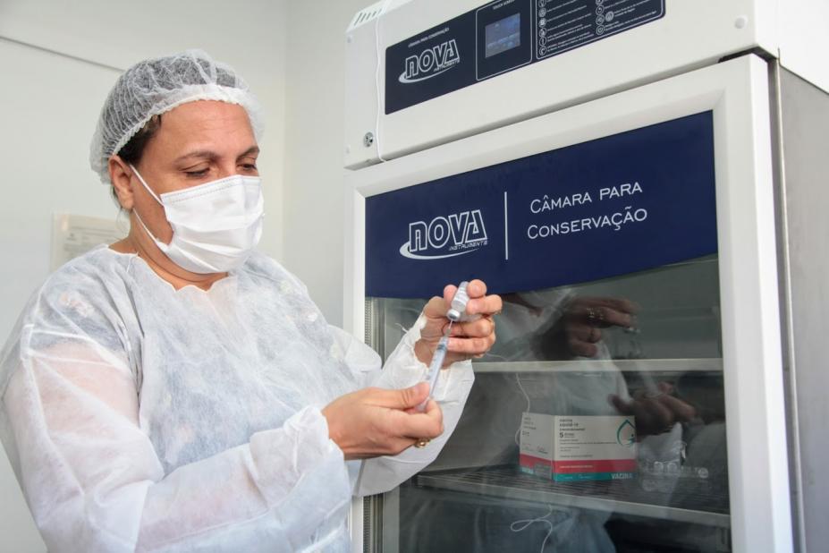 mulher está colocando o imunizante em seringa. Ela está diante de um refrigerador específico para a vacina. #paratodosverem