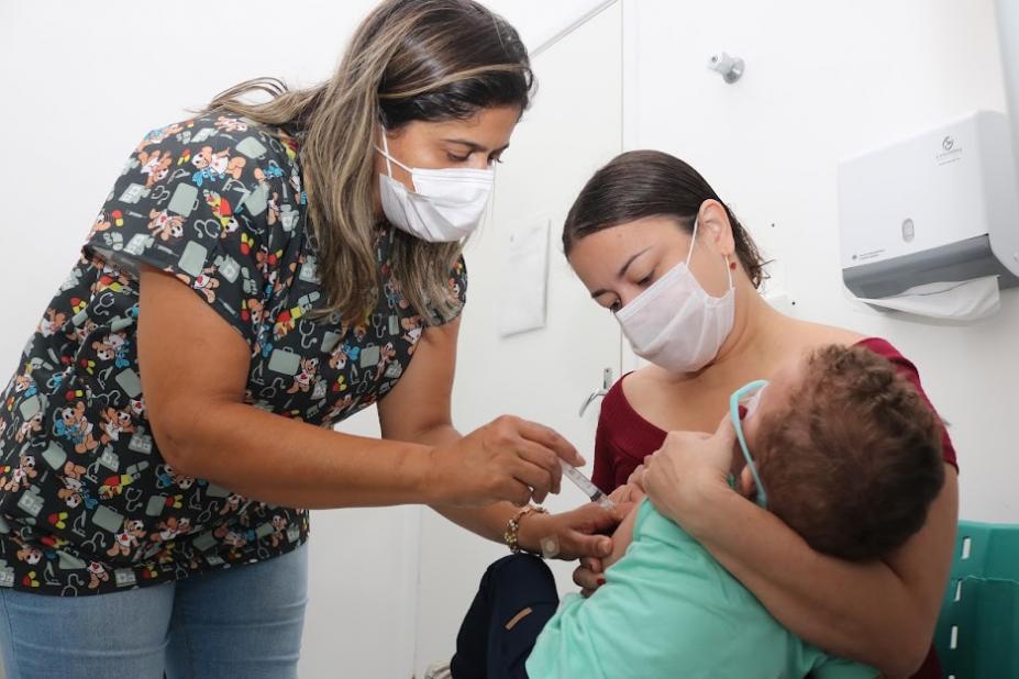 criança está no colo da mãe sendo vacinada por uma mulher. Todos estão de máscara. #paratodosverem 