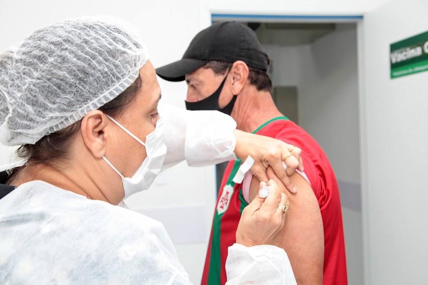 mulher usando máscara, gorro e avental vacina homem no braço esquerdo. #paratodosverem