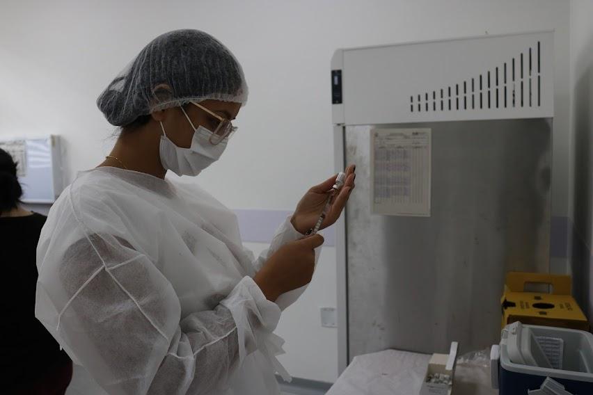 mulher usando touca, máscara e avental prepara vacina. Ela está ao lado de uma geladeira e à frente de uma mesa. #paratodosverem