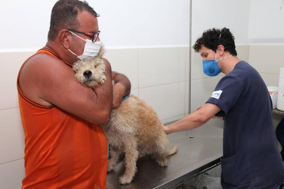 HOmem abraça cachorro que é preparado para receber vacina. Vacinador está usando máscara e aproxima a mão esquerda do animal. #Paratodosverem