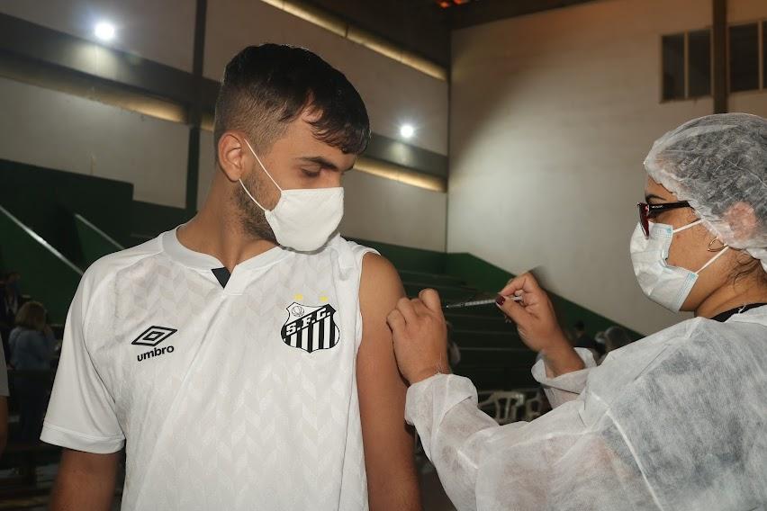 Jovem, com a camisa do Santos, recebe aplicação de vacina no braço por enfermeira paramentada. #pracegover