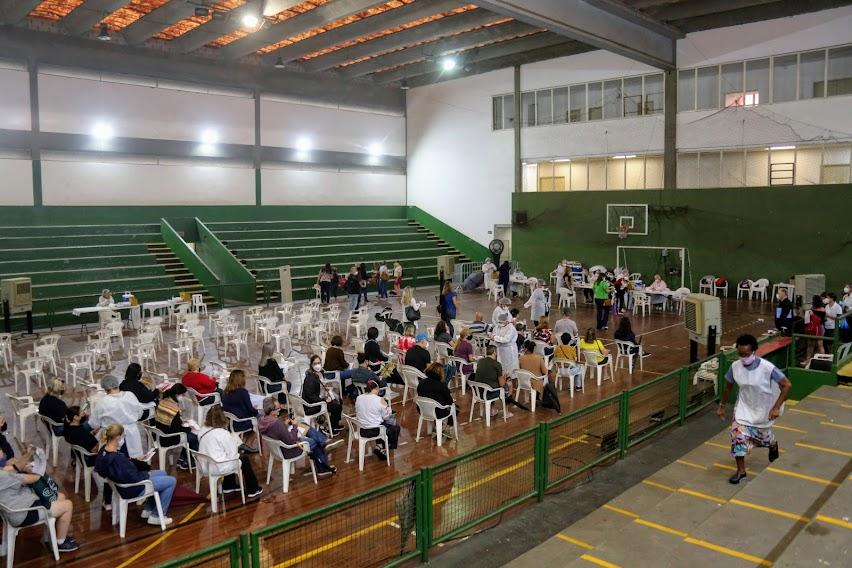 Em um ginásio, várias pessoas sentadas na cadeira aguardando vacinação. #paratodosverem