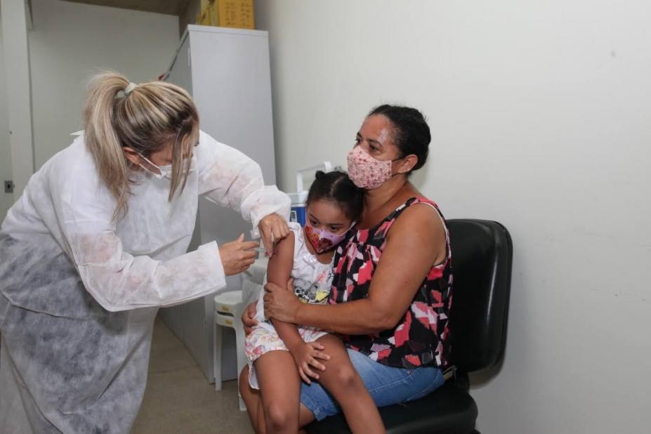 Mulher aplica vacina em criança sentada no colo da mãe #paratodosverem