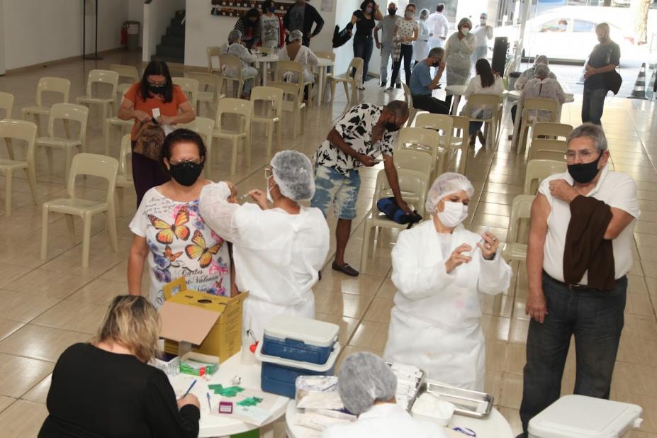 salão com várias cadeiras e pessoas em pé sendo vacinadas. #paratodosverem
