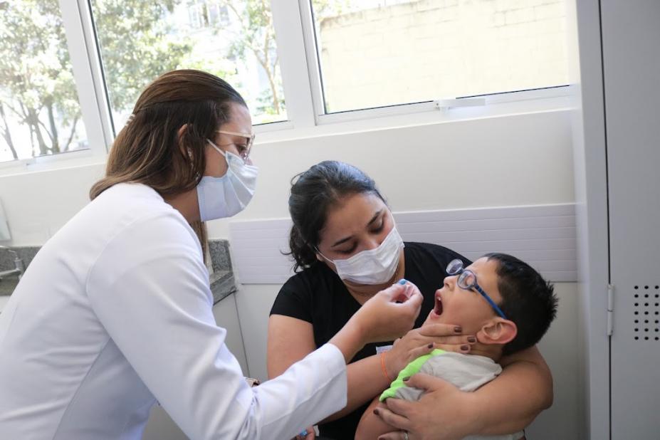 Criança, no colo do pai, recebe dose de vacina dada por enfermeira. #paratodosverem