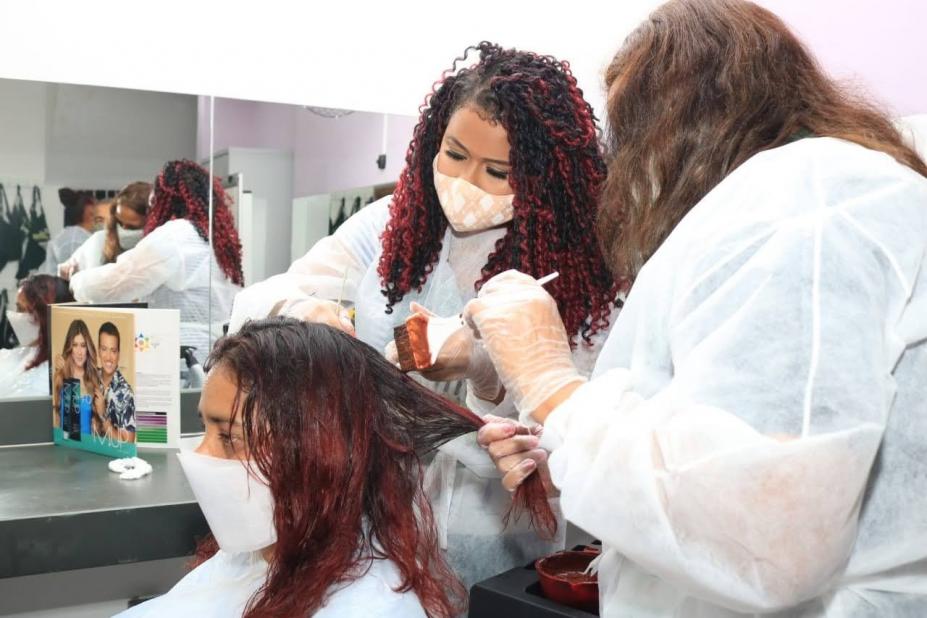 duas mulheres estão em pé pintando o cabelo de uma outra que está sentada. Há um espelho no fundo. #paratodosverem