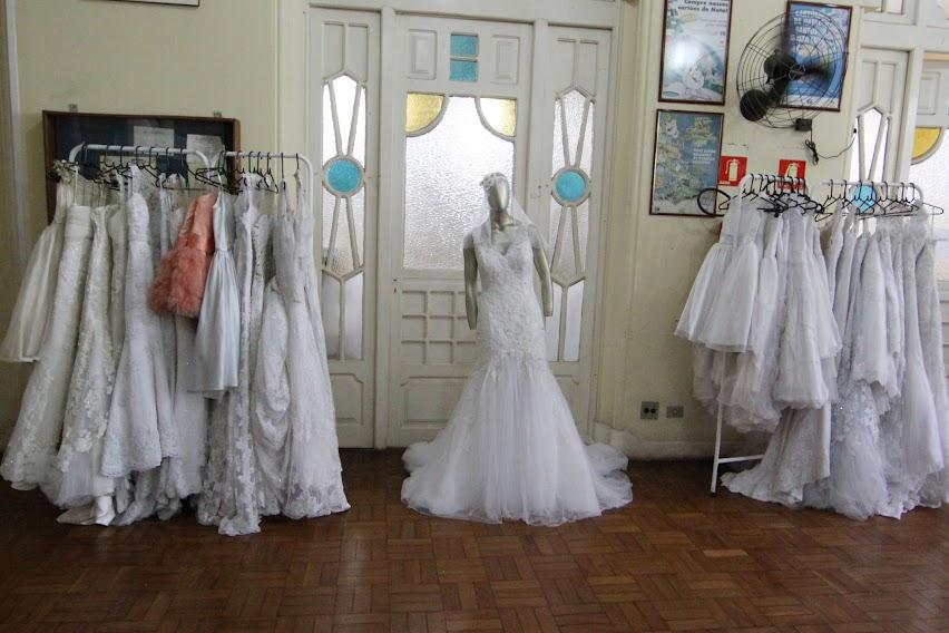 vestidos de noiva e de damas de honra estão pendurados em duas araras. Ao centro, um vestido em um manequim. #paratodosverem