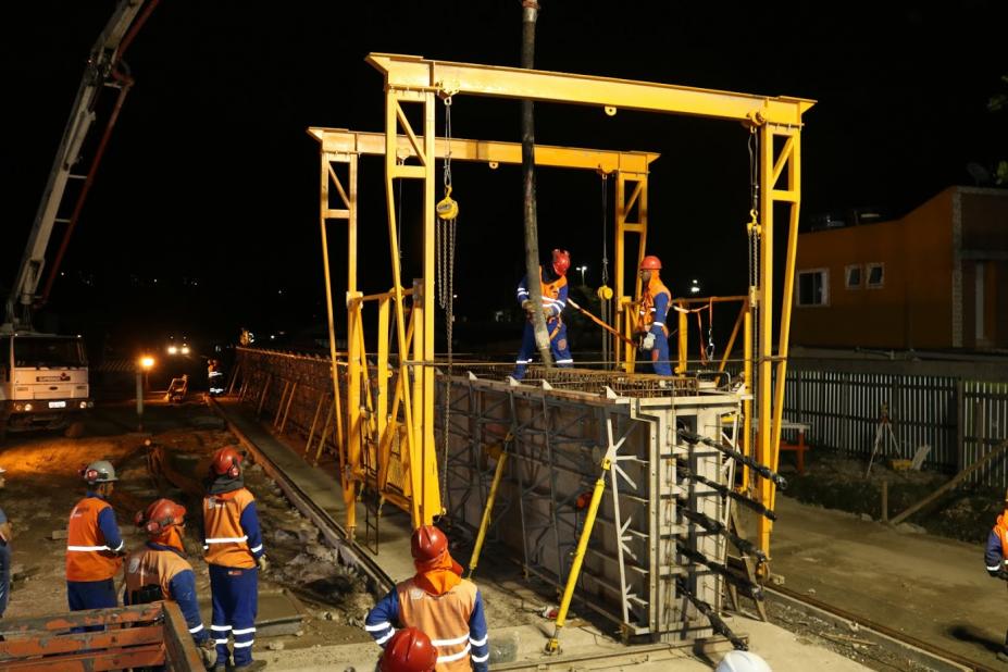 Viga está sobre o chão. Dois homens estão sobre a estrutura e seguram uma grande mangueira por onde sai o jato de concreto para preencher a haste. #Pracegover