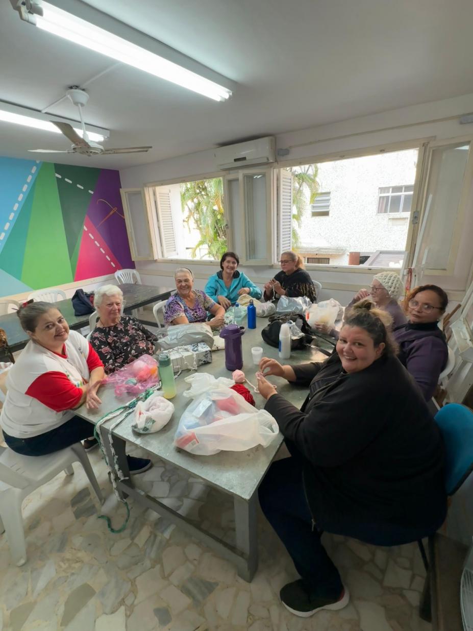 mulheres estão reunidas em torno de mesa fazendo artesanato. Algumas olham para a foto e sorriem. #paratodosverem 