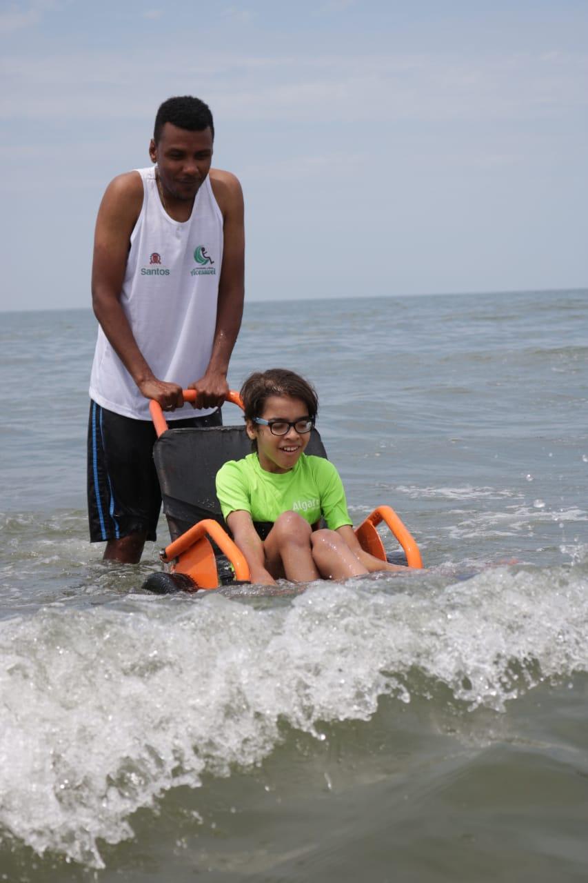 Criança toma banho de mar com ajuda de cadeira anfíbia e monitor. #pracegover