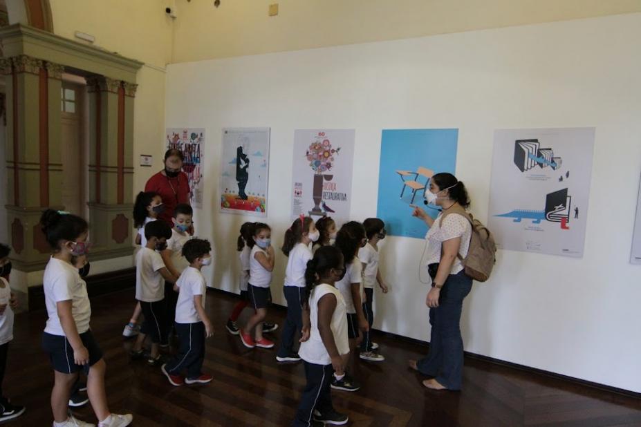 alunos estão dentro de sala da pinacoteca onde se vê cinco obras afixadas em parede. Uma mulher aponta para uma das obras enquanto as crianças olham. #paratodosverem