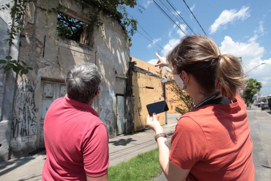 Duas pessoas estão diante de um imóvel. Ambas estão de costas para a imagem. Uma delas fotografa o imóvel com smartphone. #Paratodosverem