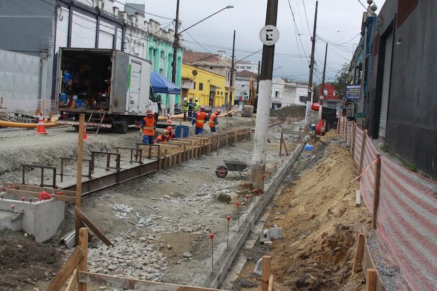 homens trabalham em obra na rua. A rua está sem asfalto. Há estruturas de madeira montadas. Um caminhão ao fundo. #paratodosverem