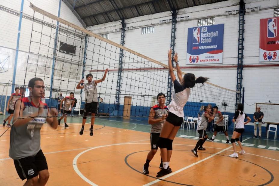 jovens estão na quadra jogando vôlei. A rede aparece no meio com alunos de cada um dos lados. #paratodosverem