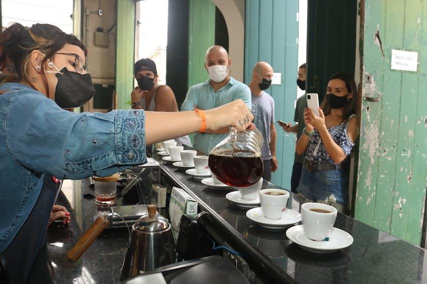 Barista serve café para grupo de turistas, que observam e tiram fotos. #pracegover