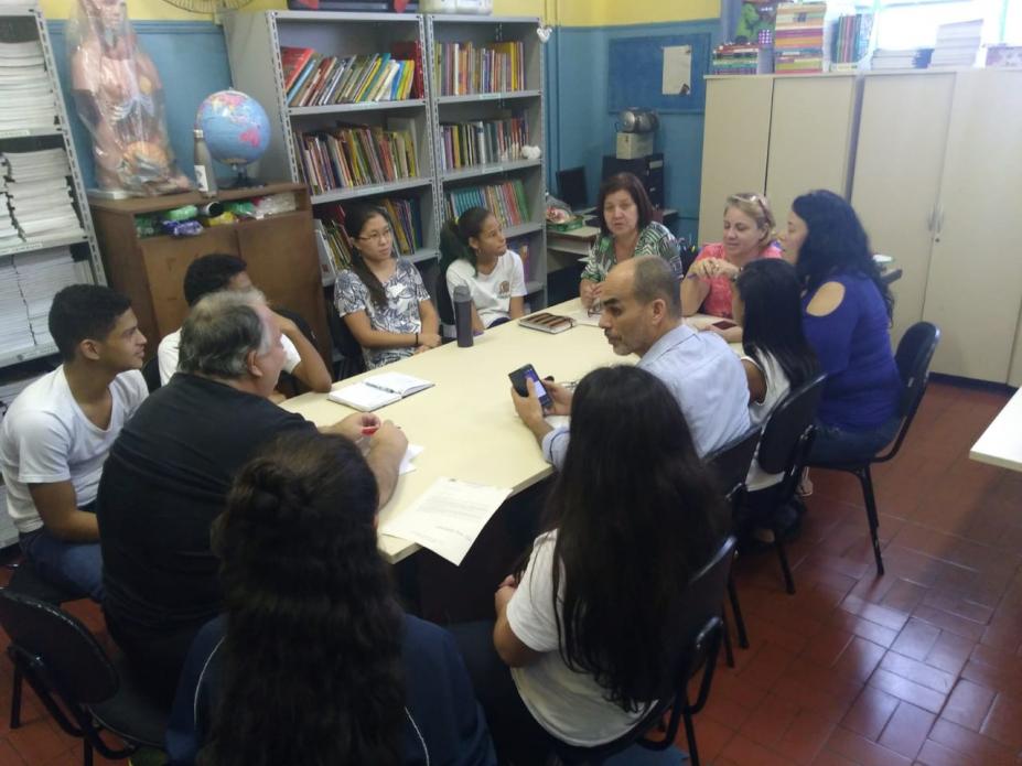 Representantes de secretarias, da escola e alunos estão sentados em torno de uma mesa. Em uma das paredes está encostada uma estante de livros. #Pracegover