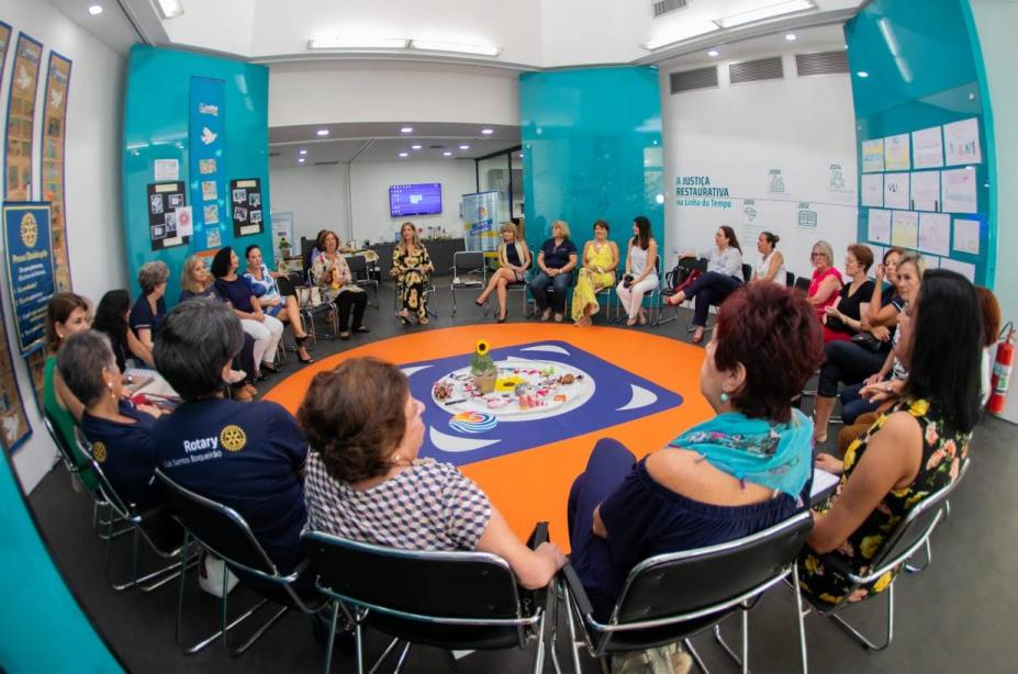 Várias pessoas estão reunidas em um círculo. Há uma mesa ao centro com o bastão que será passado entre os integrantes do círculo. #Pracegover