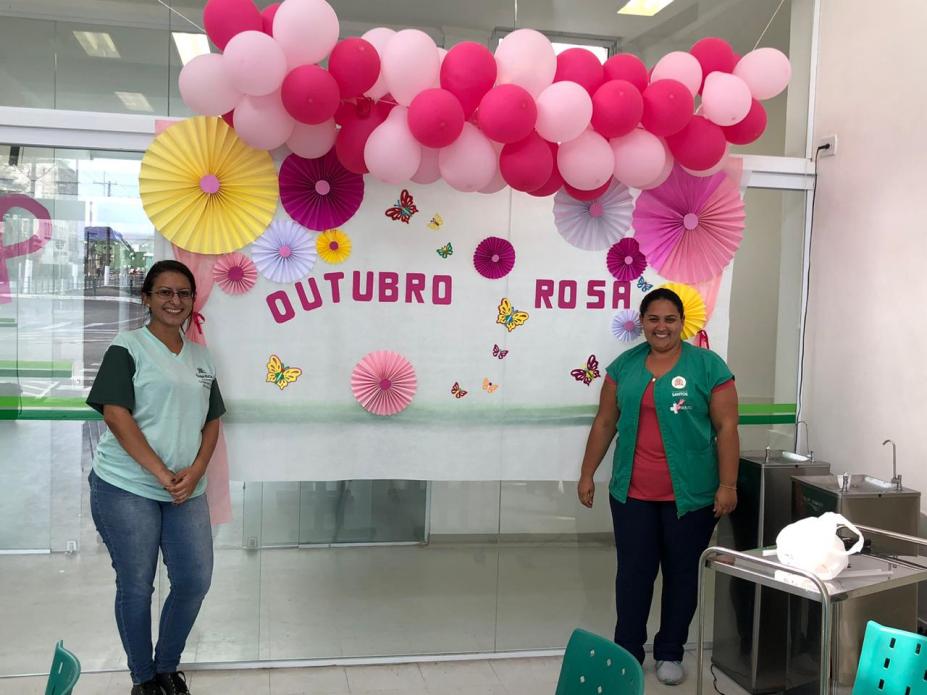 Funcionárias de policlínica estão diante de um painel com balões de gás cor de rosa. Ao fundo se lê Outubro Rosa. #Pracegover