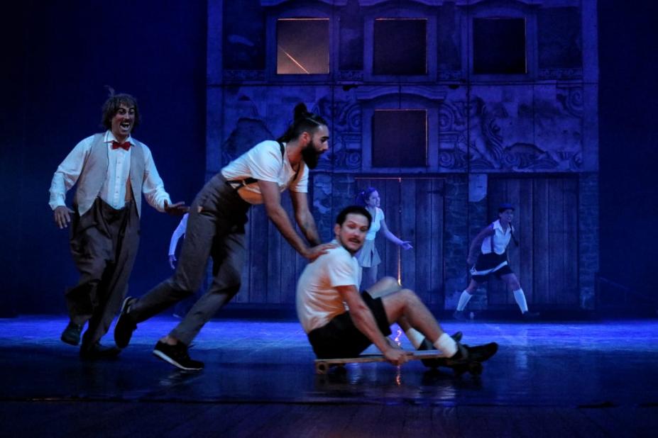 Homem empurra outro sobre skate sobre palco com luz azul