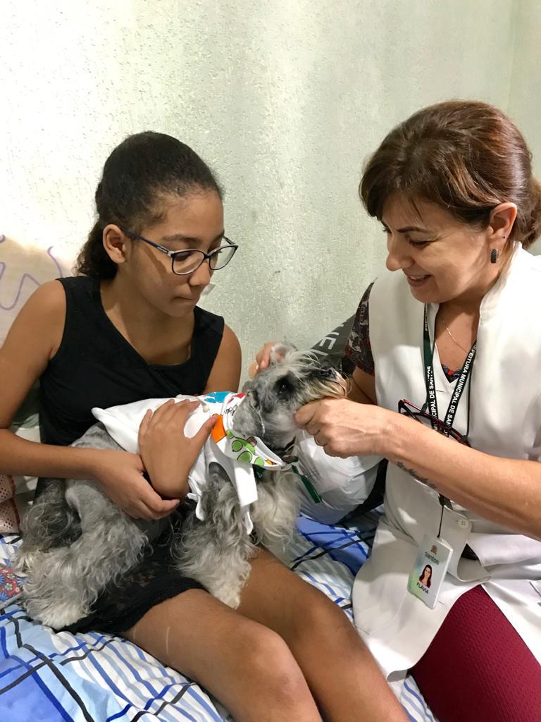 Menina segura cão em seu colo. Ao lado dela está uma mulher que usa um avental branco. #Pracegover