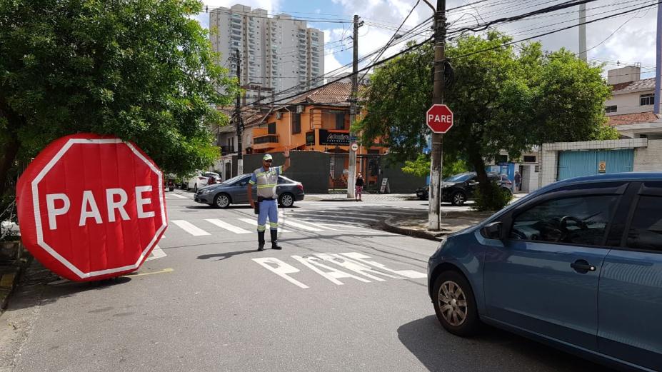 Inflável de dois metros está em primeiro plano, à esquerda, próximo a cruzamento de ruas. Ao lado direito dele, um agente de trânsito e um carro em sua direção. #Paratodosverem