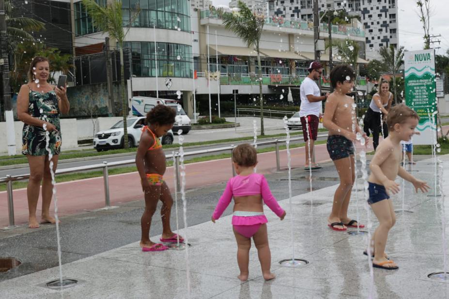 #pracegover Abservadas por três adultos, duas meninas e dois meninos brincam sobre a fonte interativa