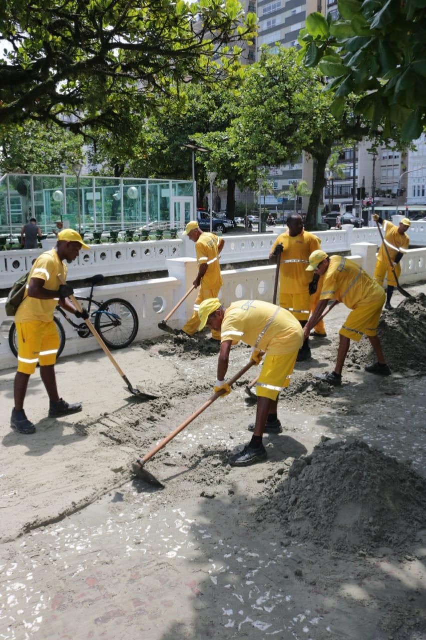 #pracegover Junto a mureta de canal na orla da praia, seis homens uniformizados de amarelo removem areia com pás
