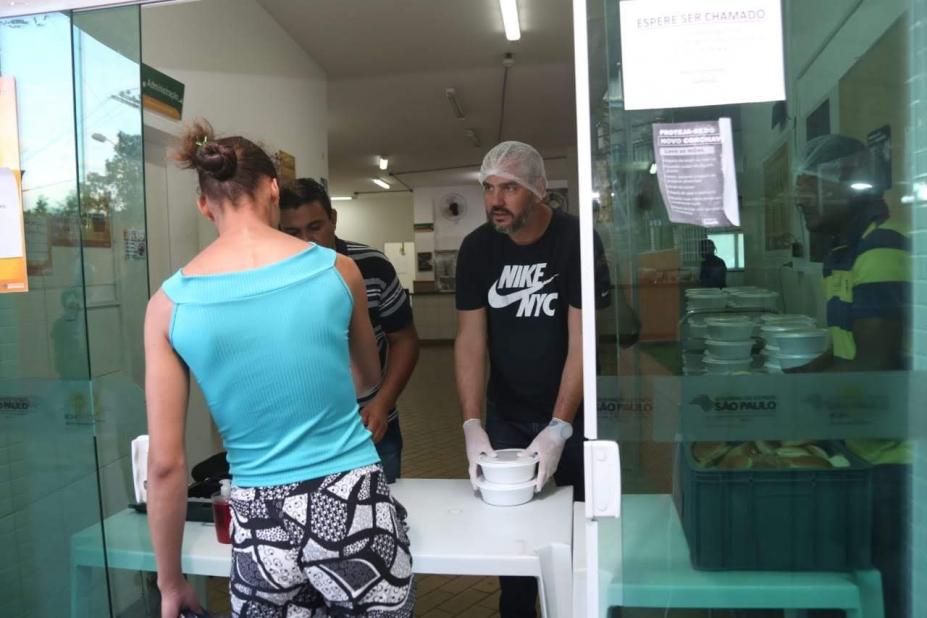 Mulher está de costas pegando refeição em embalagem que é entregue por dois homens atrás de um balcão. Eles estão atrás de uma porta de vidro. #Paratodosverem