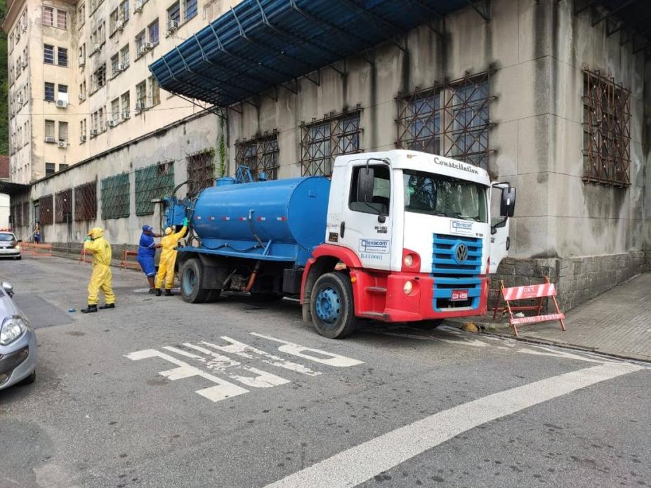 Caminhão com hidrojato está parado em rua ao lado de imóvel. Homens estão manuseando máquina na parte de trás do veículo. #Paratodosverem