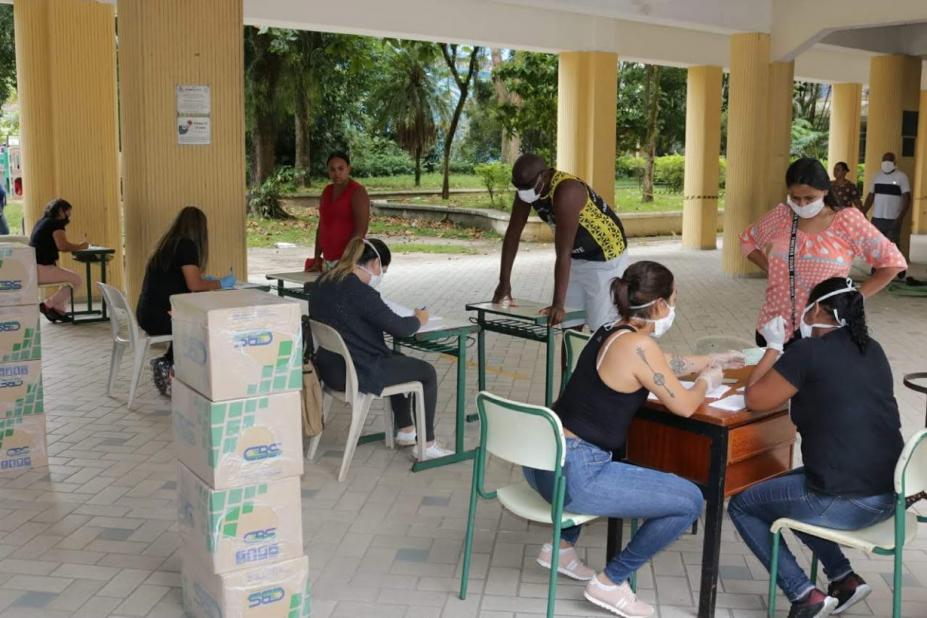 Pilhas de cestas basicas dispostas em salão. À frente delas estão três estações de atendimento ao público. Três mulheres estão sentadas atendendo o público. As pessoas usam máscaras e estão distantes umas das outras. #Paratodosverem