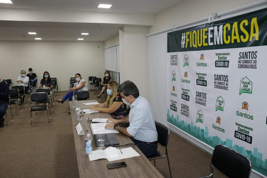 Sala com um painel à direita na parede onde se lê Fique em Casa e vários logotipos municipais abaixo. Em uma mesa estão sentadas duas pessoas usando máscaras. Ao fundo há quatro pessoas sentadas em várias cadeiras disponíveis. #Paratodosverem