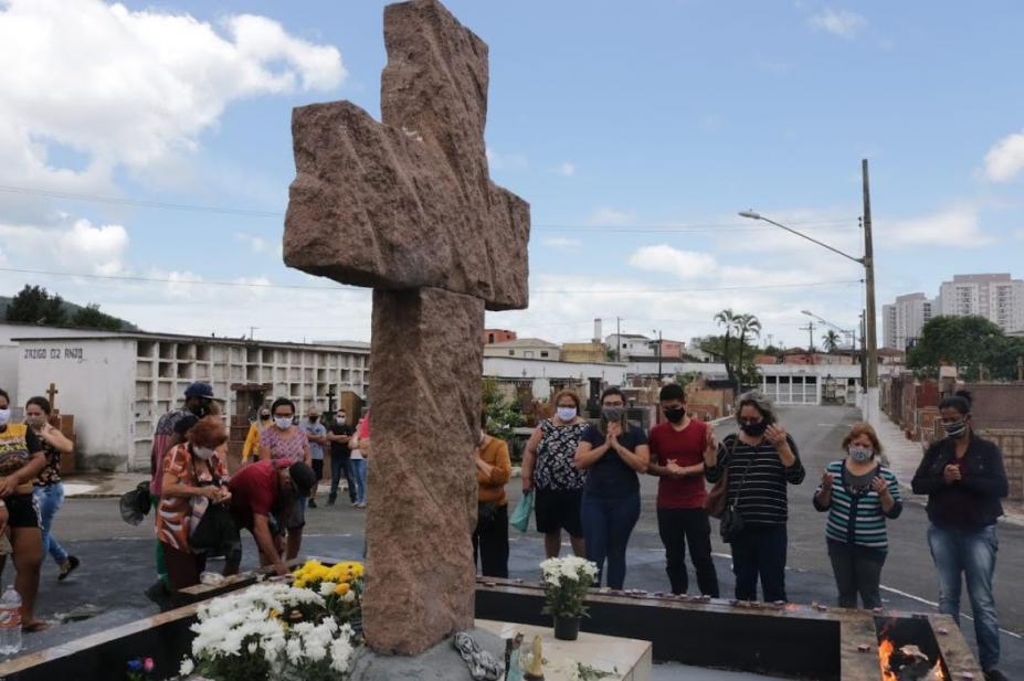 Pessoas rezando em direção a monumento de uma cruz no cemitério #paratodosverem