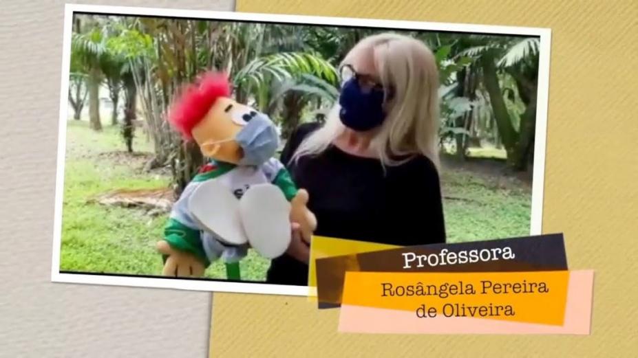 em  trecho do vídeo, professora segura fantoche, ambos de máscara #paratodosverem 