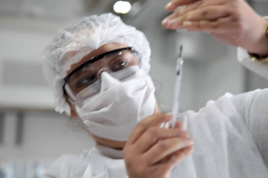 mulher usando máscara prepara vacina com agulha na mão. #paratodosverem