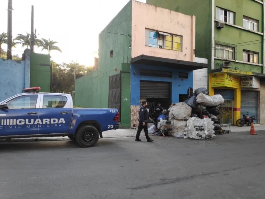 Em uma rua, viatura da guarda, agente andando e sacos com sucata em frente ao prédio. #paratodosverem