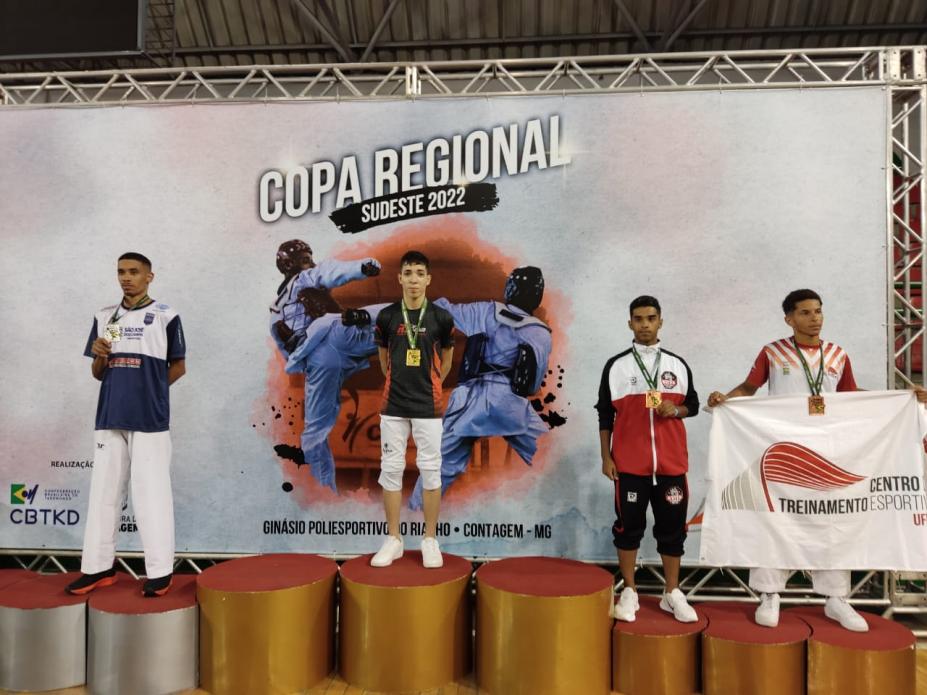 quatro atletas de pé em um pódio , o da direita tem uma bandeira aberta #paratodosverem