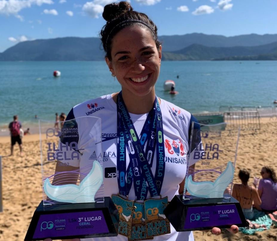 atleta na praia segurando troféus e medalhas #paratodosverem