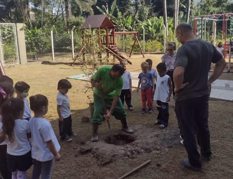 funcionário planta árvore em jardim da escola com várias crianças observando #paratodosverem